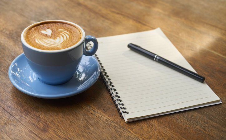 Blue coffee cup next to notepad and pen on a wooden table
