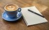 Blue coffee cup next to notepad and pen on a wooden table